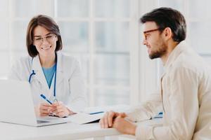 Horizontal shot of doctor and patient discuss something, talk about diagnosis, use laptop computer, pose in consulting room, check medical records. Sick man gets prescription from therapist. photo