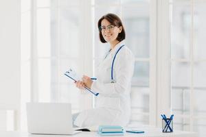 Woman doctor sits on table, writes down information in clipboard, poses at workplace in hospital office, works with laptop computer, smiles pleasantly. Health, technology, medicine, internet concept photo