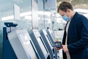 Sideways shot of man poses at self check in desk in airport, holds passport, going to register for flight, wears medical mask for safe traveling during coronavirus outbreak. Pandemic in public place photo