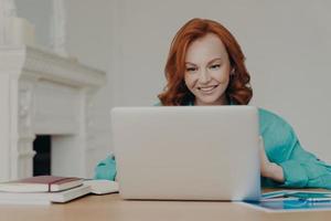 Focused cheerful redhead successful businesswoman does research, makes business project, sits in front of laptop computer at home office desk, uses software application, has happy smile on face photo