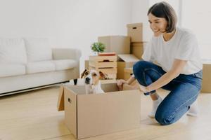 Indoor shot of pleased brunette woman dressed in white t shirt and jeans, plays with pedigree dog, moves carton box with animal, pose in living room, buy first new property. Mortgage concept photo