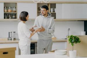 Photo of happy husband and wife unpack things after moving into new house, carry white plates, pose against kitchen interior, have fun, dressed in domestic clothes. People and moving concept