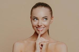 Portrait of good looking woman looks aside mysteriously, keeps finger on chin and smiles broadly, looks away and shares secret of beauty, has perfect glowing skin, naked body, natural makeup photo