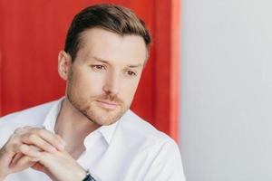 Photo of pensive male entrepreneur looks thoughtfully aside, keeps hands tgether, dressed in elegant white shirt, thinks about climbing on career ladder. People, business and thoughts concept
