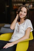 Vertical shot of pretty girl dressed in white t shirt and jeans, has happy look at camera, sits at yellow armchair, being in good mood, feels carefree, has pleasant smile on face. Feminity and rest photo