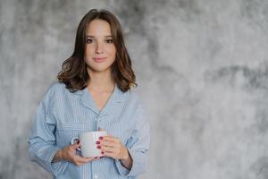 Attractive woman dressed in nightwear, holds white mug with coffee or tea, has morning drink, poses indoor against blurred background with copy space for your advertisement or promotional content photo