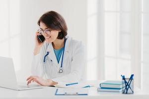 Online consultation concept. Cheerful female medical worker talks via cellular, searches useful information in internet, works on laptop computer, sits at desktop with notebooks, clipboard, pens photo