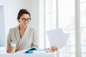 Horizontal view of confident brunette female worker in formal wear and optical spectacles looks through documentation, drink fresh beverage, focused down, poses indoor, has pleased expression photo