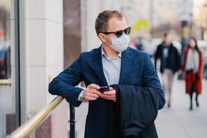 Prosperous businessman in formal wear poses at street, waits for someone, holds mobile phone and sends text messages, wears medical mask during coronavirus outbreak, few people walking outside photo