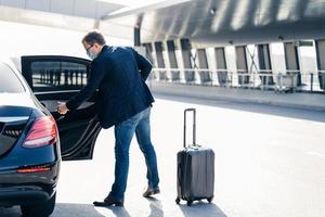 Man arrives from abroad, wears medical mask, returns from country with coronavirus outbreak, needs quarantine and self isolation, opens doos of car, places luggage in transport. Virus spread concept photo