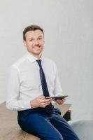 Vertical shot of happy businessman in formal clothes checks email and reads notification on modern tablet computer, sit at wooden desk in office building. People, business and technology concept photo