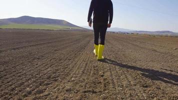 granjero caminando en el campo. granjero moderno que sostiene una tableta camina con botas amarillas en una tierra de cultivo. mira a su alrededor y examina el terreno. video
