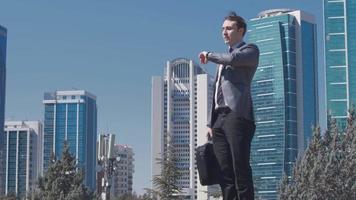 empresario nervioso llegando tarde, edificios comerciales. el hombre de negocios de traje con el bolso en la mano mira su reloj y sigue su camino nervioso porque llega tarde. video