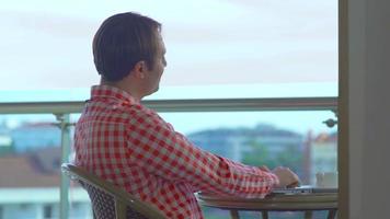 empresario bebiendo café en el balcón del hotel. hombre sentado en el balcón del hotel con vista al mar y sosteniendo una computadora portátil frente a ella bebiendo su café. video