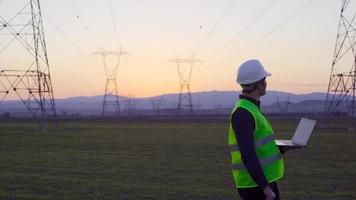 Electrical engineer working in front of high voltage lines. Engineer working on laptop at sunset and looking at electric poles. Field research and review video