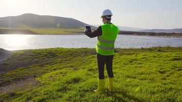 ingeniero haciendo estudios de tierra y agua. el ingeniero, sosteniendo una tableta en el campo, realiza investigaciones alrededor de la tierra y el lago. video