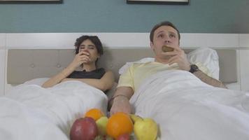 le couple mange des fruits au lit et regarde la télévision. le couple avec des fruits dans les mains regarde la télé dans sa chambre. video