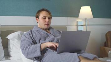 Mann, der am Laptop arbeitet, während er im Bett liegt. mann liegt im bademantel auf dem bett, arbeitet am laptop und trinkt seinen kaffee. video