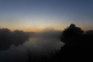 Foggy morning on a European river with fresh green grass in the sun. The rays of the sun through the tree. photo