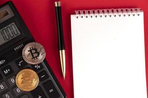 bloc de notas blanco con calculadora, bolígrafo elegante y monedas de bitcoin en un fondo rojo. copie el espacio foto