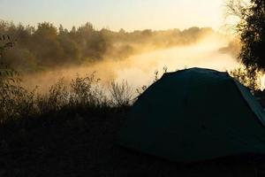 Green tourist tent by the river at sunrise, with morning autumn fog on the water. Outdoor tourist landscape. photo