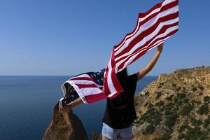 Rear view of a young woman holding an American flag waving at the coastline against the sunny bright sea. photo