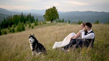 The newlyweds and their husky are sitting in the grass on a background of mountains.