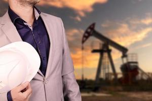 Petroleum engineer working in the oil industry stands with a white helmet in his hands in front of an oil pump. photo