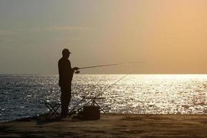 un pescador está pescando con una caña de pescar en la orilla del mar, frente al sol. foto