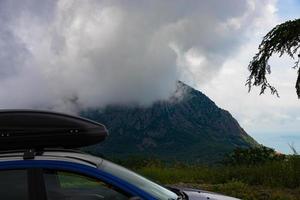 A blue car with a black roof rack stands against the backdrop of a mountain lake with mountains and clouds. photo