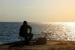 un pescador está pescando con una caña de pescar en la orilla del mar, frente al sol. foto
