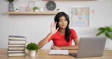 Portrait of Asian schoolgirl studying online making notes in copybook and waving hand via laptop at home. Distance learning and education concept. video