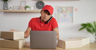 Portrait of Young Asian busy postman in red uniform and cap sitting at desk and talking on mobile phone in postal office store and working at laptop. Parcels front side. video