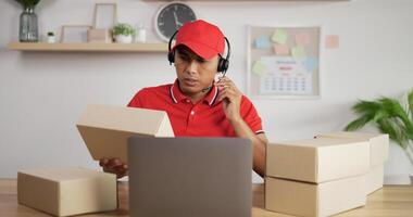 portret van jonge Aziatische drukke postbode in rood uniform, koptelefoon en pet zittend aan een bureau en praten met de klant in de postkantoor winkel en kijken naar klembord. pakketten voorzijde. video