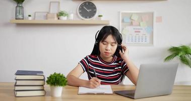 Portrait of Young Asian customer service support agent telemarketer wearing headset looking at laptop make business conference internet video call. Woman making note.