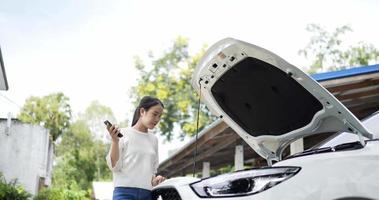 Asian woman whose car broke down is calling a mechanic. Young female standing at broken car searching for repair service on smartphone worrying in stress. Car service concept.