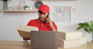 Portrait of Young Asian postman in red uniform and cap sitting at desk and talking on mobile phone in postal office store and working at laptop.