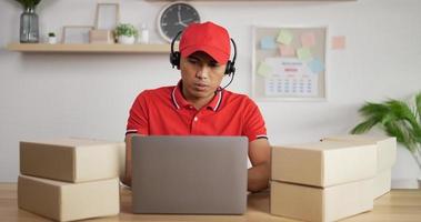retrato de jovem carteiro asiático de uniforme vermelho, fone de ouvido e boné sentado na mesa na loja postal e trabalhando no laptop.