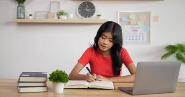 porträt eines asiatischen schulmädchens, das online lernt und notizen in einem schreibheft mit laptop auf dem tisch zu hause macht. Fernlernkonzept. video