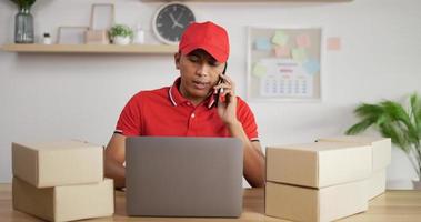 Portrait of Young Asian busy postman in red uniform and cap sitting at desk and talking on mobile phone in postal office store and working at laptop. Parcels front side.