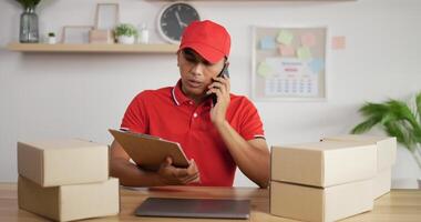 porträtt av ung asiatisk upptagen brevbärare i röd uniform och mössa som sitter vid skrivbordet och pratar i mobiltelefon i postkontorets butik och tittar på urklipp. paket framsidan. video