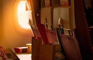 Food in brown paper bag put on plastic airplane tray table at seat back with blurred passenger hand opened black leather bag. Sunlight passing through the economy class airplane window into the plane. photo