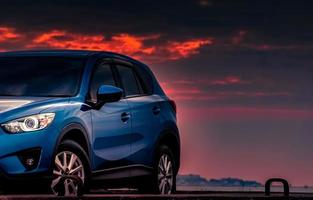 New blue SUV car with sport and luxury design parked on concrete road by the sea at sunset with dramatic sky and clouds. Automotive industry. Hybrid and electric car technology. Summer travel in night photo