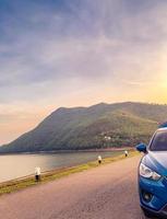 Front view of blue SUV car with opened car truck parked on asphalt road at dam. Summer road trip travel. Car parked on the road in front of the mountain beside reservoir. Summer vacation with nature. photo