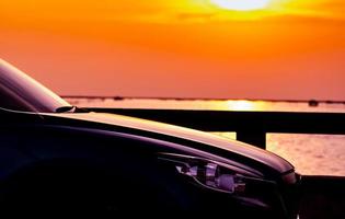 Side view of SUV car with sport and modern design parked on concrete road by sea beach at sunset. Hybrid and electric car technology. Road trip travel. Automotive industry. Orange and yellow sky. photo
