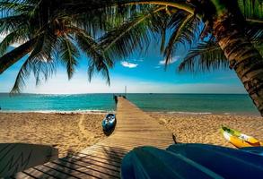 Beautiful view tropical paradise beach of resort. Coconut tree, wooden bridge, and kayak at resort on sunny day. Summer vacation concept. Summer vibes. Golden sand beach of resort with blue sky. photo