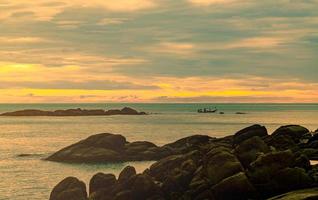 Beautiful stone beach in the morning with golden sunrise sky. Fisherman in long tail boat with folk fishing culture. Peaceful and tranquil scene. Calm sea in the morning. Seascape with skyline.