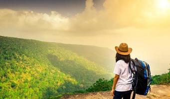 Young traveling woman with backpack and hat stand on the top of the mountain cliff watching beautiful view of woods and sky after rain on her vacation. Asian woman travel alone. photo