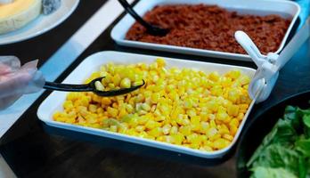 Selective focus on sweet corn in white bowl at salad bar buffet of restaurant. Hand wear plastic gloves holding black spoon scoop yellow sweet corn. Healthy eating. Healthy food. Vegetarian food. photo