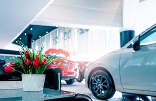 Fake red flowers in ceramic white vase on glass table in car showroom. Car dealership. Artificial flower on blurred new and shiny luxury car parked in modern showroom for sale. Showroom interior. photo